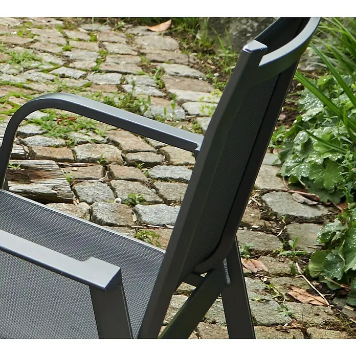 Acamp Stapelsessel Urban - 8 Stk., Aluminium, Acatex, Grau, B x T x H: 62 x 69 x 97 cm - Nahaufnahme eines grauen Gartenstuhls mit Armlehne vor einem Hintergrund aus Pflastersteinen und Grünpflanzen.