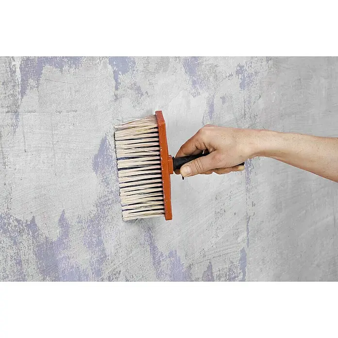 Knauf Rotband Grundierung - 1 kg - Eine Hand hält eine Quastbürste, um eine unebene graue Wand zu grundieren, wobei violette Farbreste sichtbar sind, die eine vorbereitende Basis bieten.
