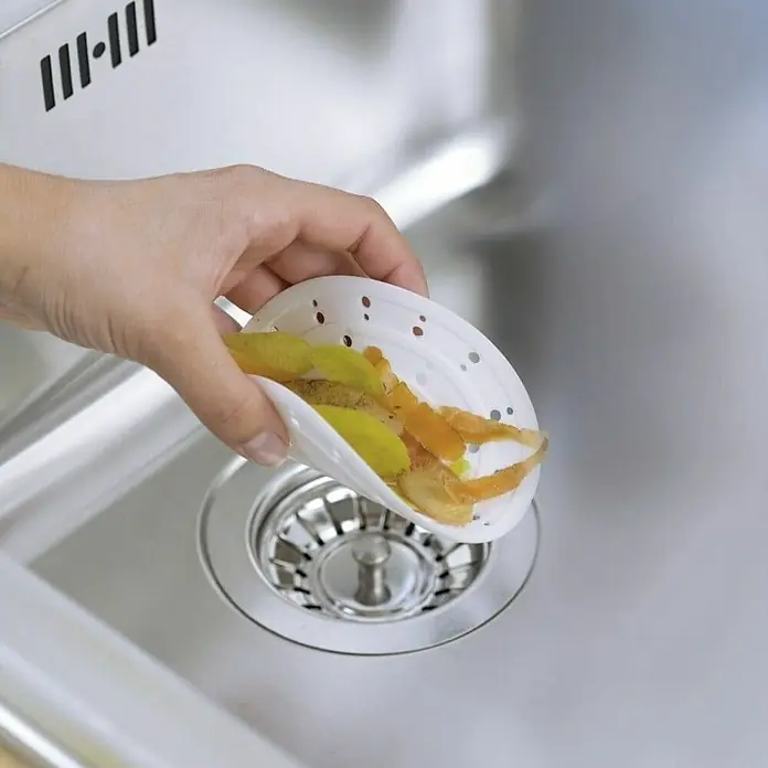 Wenko Haarfangsieb - Durchmesser: 12 cm, Silikon - Ein weißer Küchensieb mit gelben und orangefarbenen Speiseresten wird über einem Edelstahlspülbecken ausgeleert.