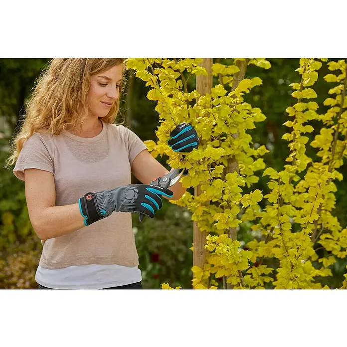 Gardena Gartenhandschuhe Strauchpflege - L, Grau/Türkis - Eine Frau mit GARDENA-Gartenhandschuhen und einer Gartenschere beim Beschneiden eines Baumes mit gelben Blättern.