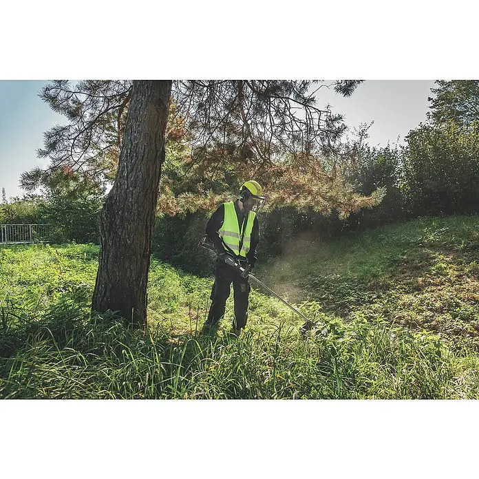 Metabo CAS 18V Akku-Rasentrimmer FSD 36-18 LTX BL 40 - 18 V, 2 Ah, Ohne Akku, Schnittbreite Faden: 40 cm - Ein Mann mit Schutzkleidung benutzt einen Rasentrimmer, um hohes Gras unter einem Baum zu schneiden, wobei der Trimmer im Fokus steht.
