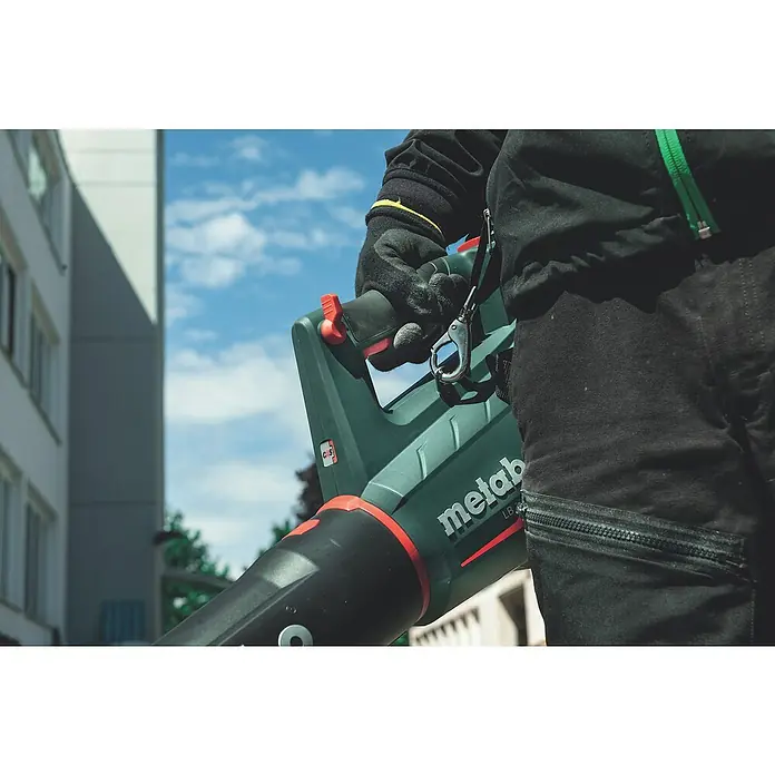 Metabo CAS 18V Akku-Laubbläser LB 18 LTX BL - 18 V, Li-Ionen, 2 Akkus, Blasgeschwindigkeit: 150 km/h - Eine Nahaufnahme eines Metabo LB 36-Akku-Laubbläsers, der von einer Person mit Handschuhen gehalten wird, mit einem Gebäude und blauem Himmel im Hintergrund.