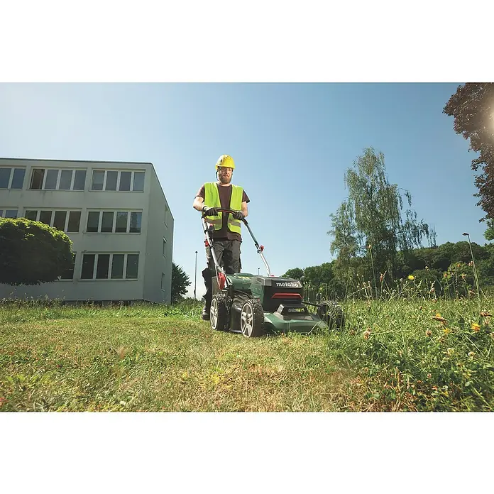 Metabo CAS 18V Akku-Rasenmäher RM 36-18 LTX BL 46 - 18 V, 2 Akkus, 5,2 Ah, Schnittbreite: 46 cm - Ein Mann mit Schutzhelm und Warnweste schiebt einen grünen Metabo-Rasenmäher über eine Wiese. Das Gerät schneidet das Gras.