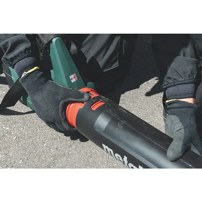 Metabo CAS 18V Akku-Laubbläser LB 18 LTX BL - 18 V, Li-Ionen, 2 Akkus, Blasgeschwindigkeit: 150 km/h - Detailaufnahme eines Metabo Laubbläsers in Aktion, der von einer Person mit Handschuhen gehalten wird. Das Gerät ist grün, rot und schwarz.