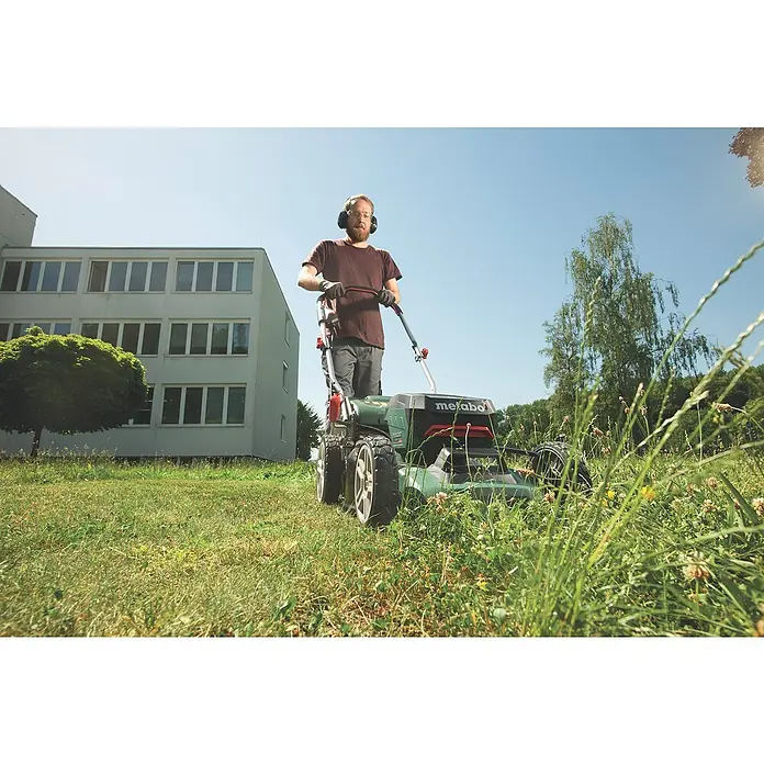 Metabo CAS 18V Akku-Rasenmäher RM 36-18 LTX BL 46 - 18 V, 2 Akkus, 5,2 Ah, Schnittbreite: 46 cm - Ein Mann mäht Rasen mit einem Metabo-Rasenmäher in einem grünen Feld, mit Gebäude und Bäumen im Hintergrund.