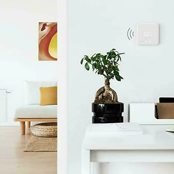Tado Sensor de temperatura inalámbrico - 1,8 x 10,4 x 10,4 cm, Blanco - Primer plano de una habitación que muestra un termostato electrónico en la pared y una planta de interior en una mesa debajo.