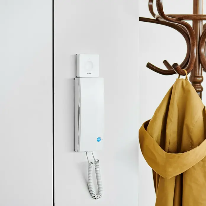 Nuki Elektrischer Türöffner Opener - Passend für: Nuki Smart Lock - Nahaufnahme eines Nuki Opener an einer weißen Wand, darunter ein Gegensprechanlage und eine beige Jacke an einem Garderobenständer.