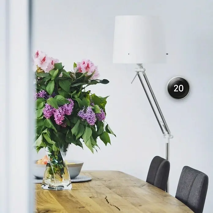Google Nest Termostato inalámbrico 3ra generación - Negro, Ø x Al: 8,4 x 8,4 cm - Termostato Nest inteligente en la pared junto a una lámpara de pie. El termostato muestra una temperatura de 20 grados, garantizando un confort óptimo.