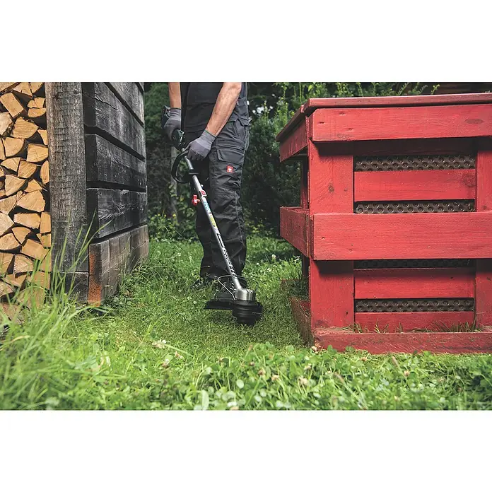 Metabo CAS 18V Akku-Rasentrimmer RTD 18 LTX BL 30 - 18 V, 2 Ah, Ohne Akku, Schnittbreite Faden: 30 cm - Ein Bediener benutzt einen Metabo Rasentrimmer, um Gras zwischen einem Holzstapel und einem roten Holzkasten zu schneiden.