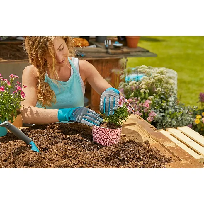 Gardena Gartenhandschuhe - M, Türkis/Grau - Eine Frau trägt Gardena Gartenhandschuhe und pflanzt Blumen in einen Topf mit Gartenerde, umgeben von Gartengeräten.