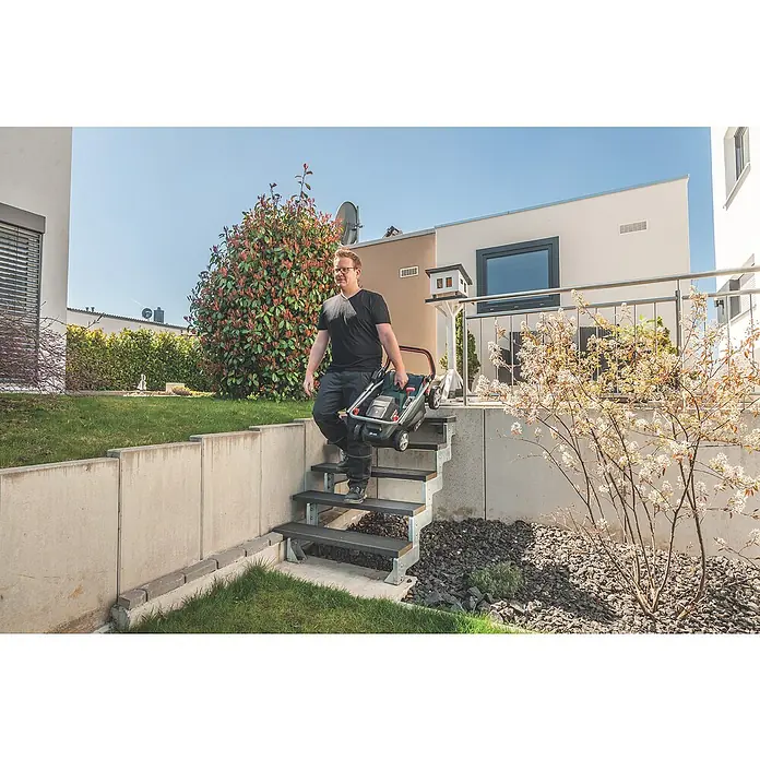 Metabo CAS 18V Akku-Rasenmäher RM 36-18 LTX BL 36 - 18 V, Ohne Akku, 5,2 Ah, Schnittbreite: 36 cm - Ein Mann trägt einen Bosch Rasenmäher die Treppe hinunter in einem Garten mit moderner Architektur und üppiger Vegetation.