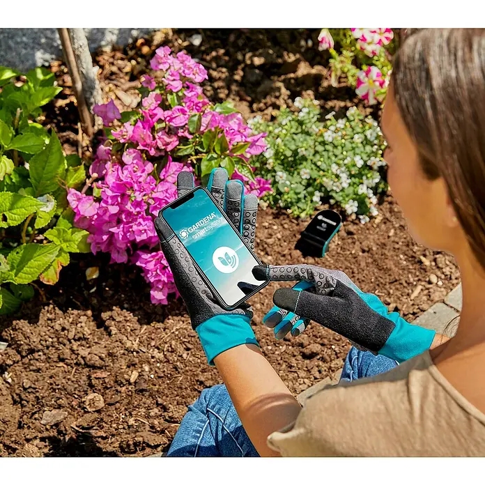 Gardena Gartenhandschuhe Geräte - L, Grau/Türkis - Frau nutzt die Gardena Smart System App auf ihrem Handy mit Gartenhandschuhen, im Hintergrund Blumen und Gartengeräte.