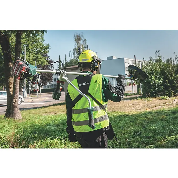 Metabo CAS 18V Akku-Rasentrimmer FSB 36-18 LTX BL 40 - 18 V, 2 Ah, Ohne Akku, Schnittbreite Faden: 40 cm - Landschaftsgärtner mit Metabo-Rasentrimmer über der Schulter. Der Trimmer ist rot und grün und der Gärtner trägt Schutzkleidung.