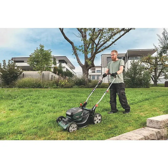 Metabo CAS 18V Akku-Rasenmäher RM 36-18 LTX BL 46 - 18 V, Ohne Akku, 5,2 Ah, Schnittbreite: 46 cm - Ein Mann mäht den Rasen mit einem Metabo Akku-Rasenmäher vor modernen Wohnhäusern. Der Mäher ist grün und schwarz, mit silbernen Rädern.