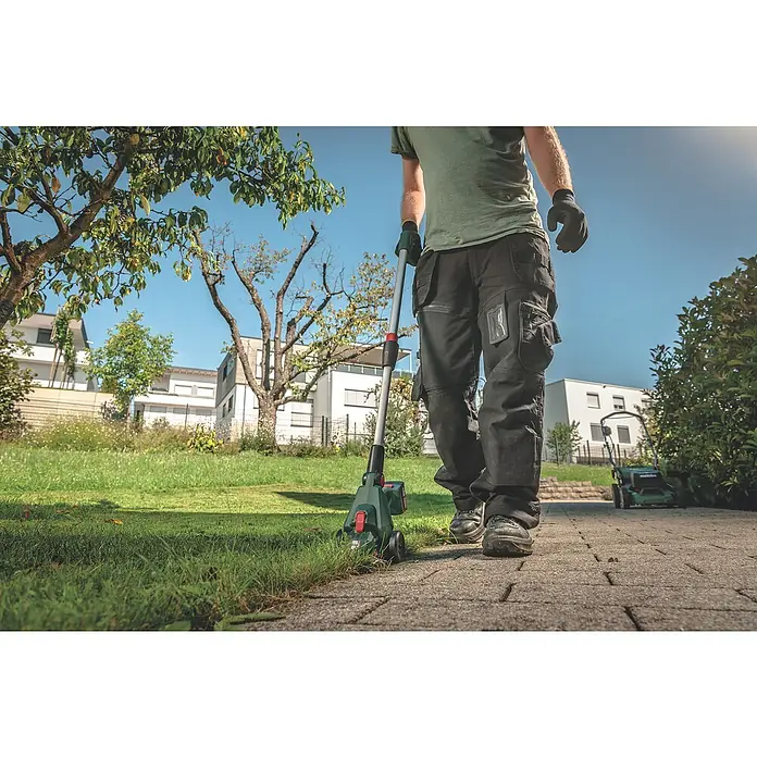 Metabo Akku-Gras- & Strauchschere PowerMaxx SGS 12 Q - 12 V, Li-Ionen, Ohne Akku, Schwertbreite: 13 cm - Mann mit Metabo Rasentrimmer mit Rad zum Rasenkantenschneiden auf gepflastertem Weg, Rasenmäher im Hintergrund.