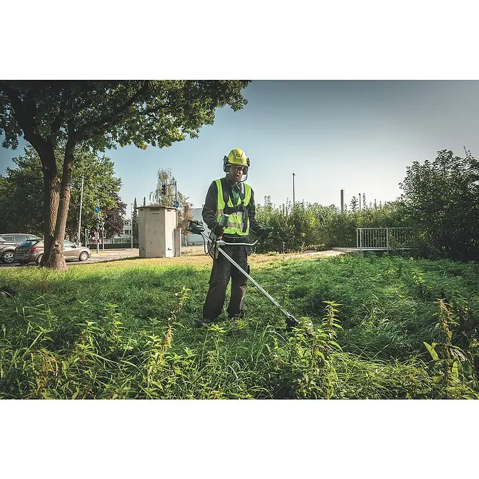 Metabo CAS 18V Akku-Rasentrimmer FSB 36-18 LTX BL 40 - 18 V, 2 Ah, Ohne Akku, Schnittbreite Faden: 40 cm - Ein Mann mit gelbem Helm und Sicherheitsweste bedient einen Freischneider in hohem Gras, umgeben von Bäumen und städtischer Umgebung.