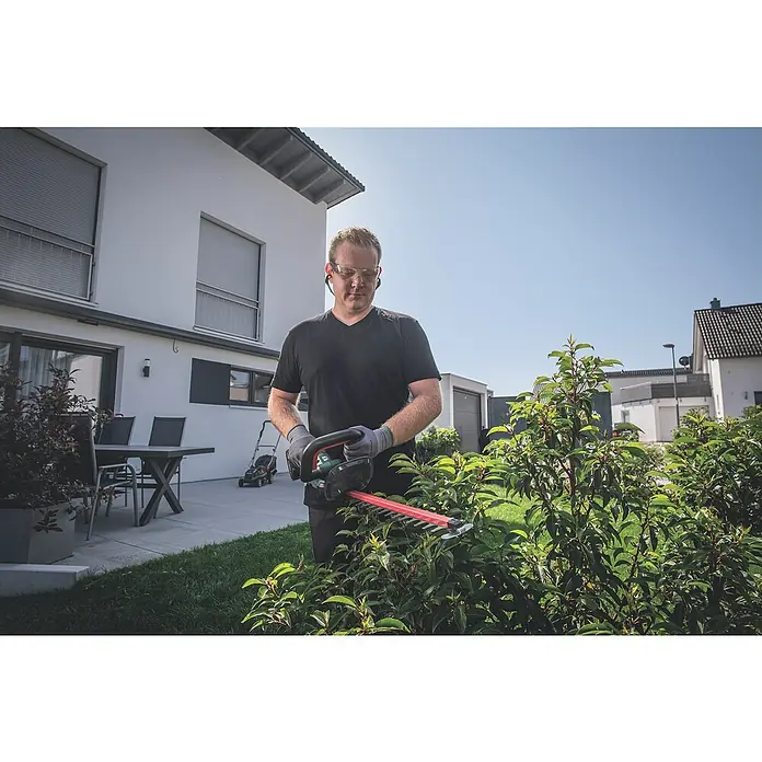 Metabo CAS 18V Akku-Heckenschere HS 18 LTX 45 - 18 V, Li-Ionen, Ohne Akku, Schwertlänge: 45 cm - Ein Mann trimmt eine Hecke mit einer roten Heckenschere von Bosch, er trägt dabei Schutzbrille und Handschuhe.
