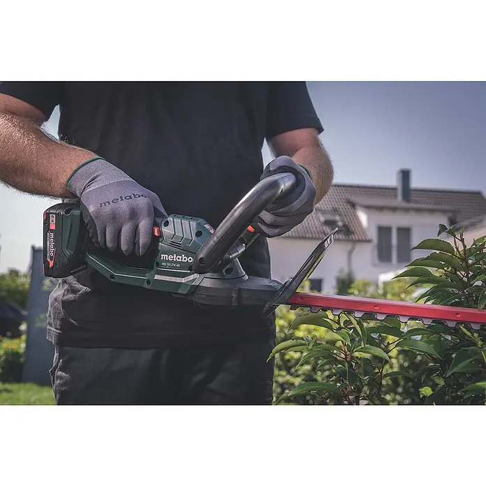 Metabo CAS 18V Akku-Heckenschere HS 18 LTX 45 - 18 V, Li-Ionen, Ohne Akku, Schwertlänge: 45 cm - Eine Person benutzt eine Metabo HS 18 LTX 45 Akku-Heckenschere mit roter Klinge und trägt graue Metabo-Handschuhe bei der Gartenarbeit.