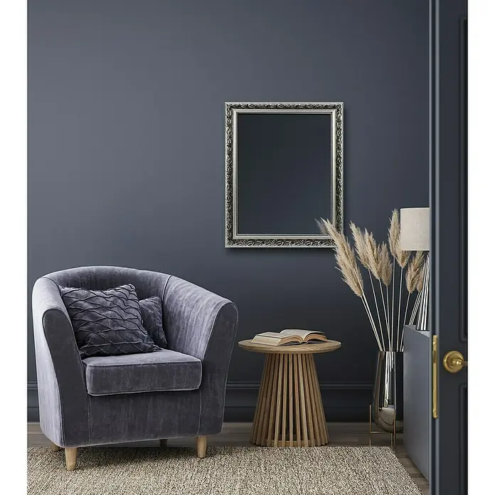 Rahmenspiegel Tanja - 55 x 70 cm, Silber, Holz - Elegantes Interieur mit einem stilvollen grauen Sessel mit Kissen, einem Beistelltisch mit Buch und einem dekorativen Spiegel an einer dunkelblauen Wand.