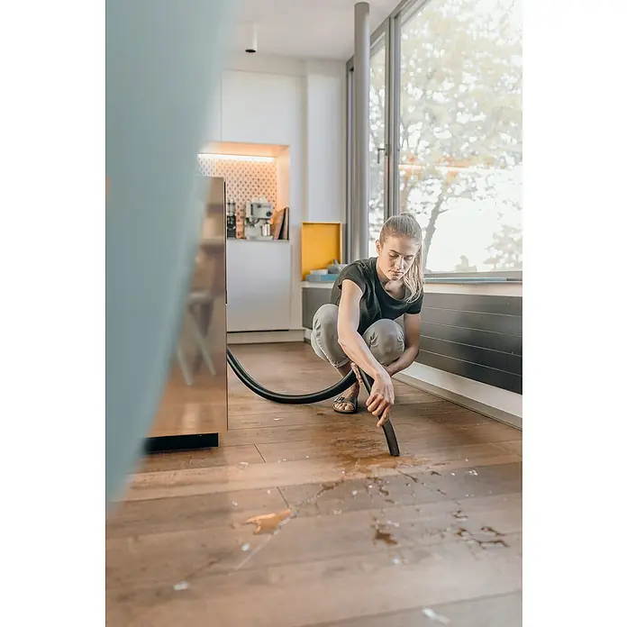 Kärcher Aspirador en seco y húmedo eléctrico WD3 Home - 1.000 W, Volumen de recipiente: 19 l, Con bolsa - Una mujer está en cuclillas en el suelo, aspirando líquido derramado de un piso de madera dura con una aspiradora.