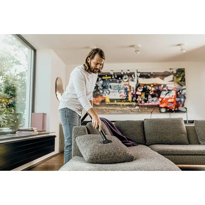 Kärcher Aspirador en seco y húmedo eléctrico WD3 Home - 1.000 W, Volumen de recipiente: 19 l, Con bolsa - Un hombre aspira un sofá de esquina gris con una aspiradora Kärcher. La atención se centra en la limpieza del sofá con la aspiradora.