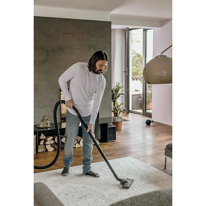 Kärcher Aspirador en seco y húmedo eléctrico WD3 Home - 1.000 W, Volumen de recipiente: 19 l, Con bolsa - Un hombre aspira una alfombra color crema en una sala de estar con pisos de madera. La aspiradora es negra y elegante.