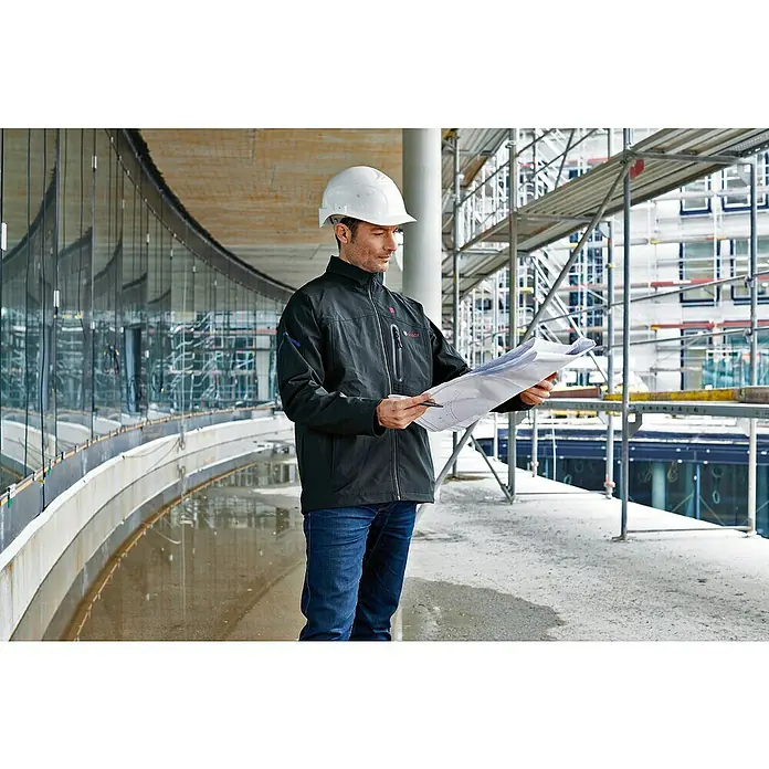 Bosch Professional Softshell-Jacke GHJ beheizbar - 2 -tlg., Akkuspannung: 12 V, M - Ein Bauarbeiter mit einem Bosch-Arbeitsjacke und einem Helm steht auf einer Baustelle und liest einen Bauplan.