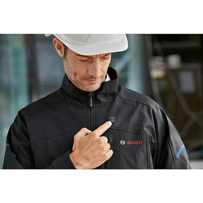Bosch Professional Softshell-Jacke GHJ beheizbar - 2 -tlg., Akkuspannung: 12 V, M - Ein Mann mit Schutzhelm präsentiert eine schwarze Bosch-Heizjacke, die Komfort und Wärme auf der Baustelle bietet.