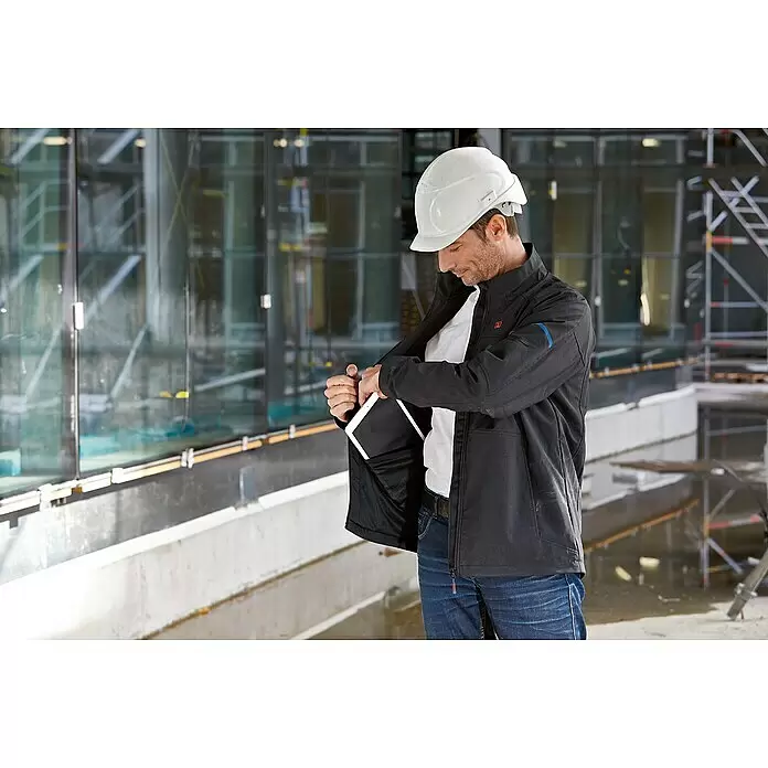 Bosch Professional Softshell-Jacke GHJ beheizbar - 2 -tlg., Akkuspannung: 12 V, M - Mann in schwarzer Arbeitsjacke mit Schutzhelm steckt Tablet in Innentasche auf Baustelle.