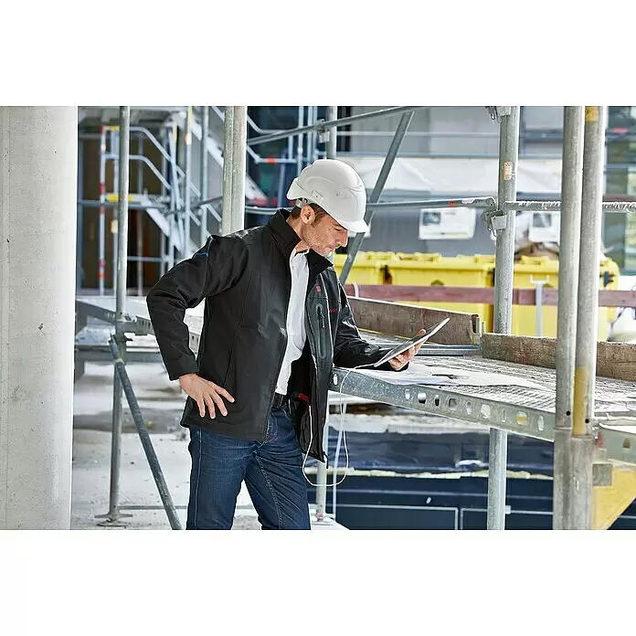 Bosch Professional Softshell-Jacke GHJ beheizbar - 2 -tlg., Akkuspannung: 12 V, M - Mann in schwarzer Arbeitsjacke und Schutzhelm prüft Pläne auf einem Tablet auf einer Baustelle.