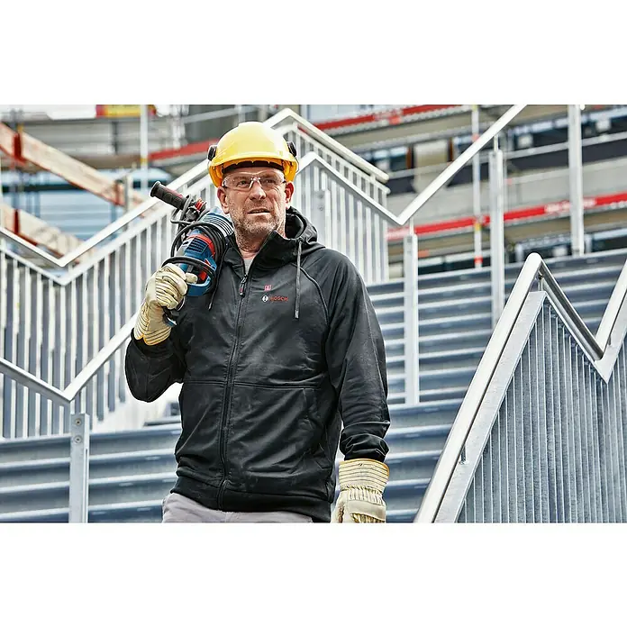 Bosch Professional Arbeitsjacke Hoodie GHH beheizbar - 2 -tlg., Akkuspannung: 12 V, XL - Ein Bauarbeiter mit gelbem Helm und Bosch-Jacke trägt eine Bosch-Bohrmaschine über einer Treppe. Sicherheitsausrüstung ist wichtig.