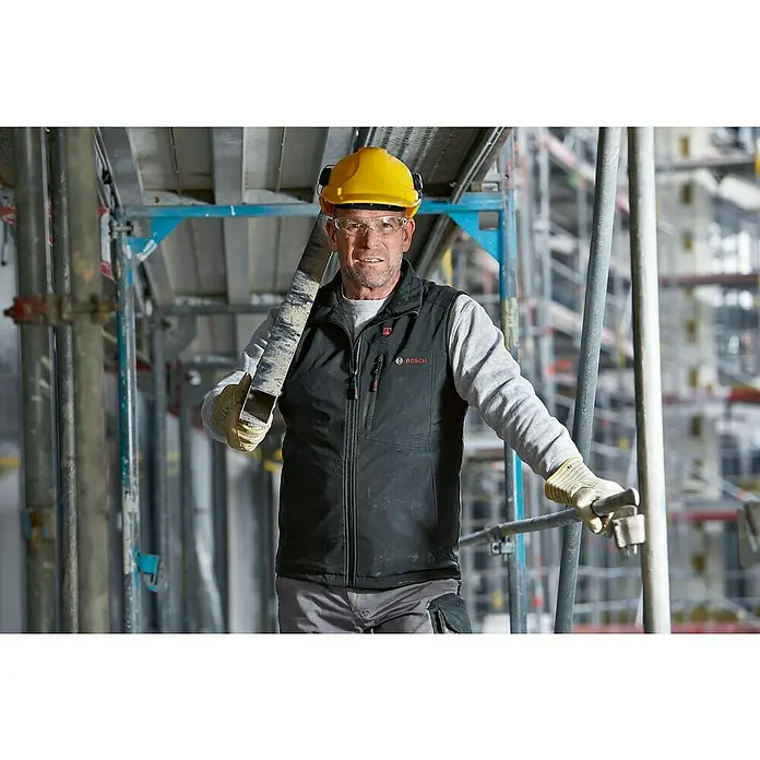 Bosch Professional Arbeitsweste GHV beheizbar - 4 -tlg., Akkuspannung: 12 V, S - Ein Bauarbeiter mit gelbem Helm und Bosch-Weste trägt ein Metallstück auf einer Baustelle. Die Bosch Weste bietet Komfort und Funktionalität.