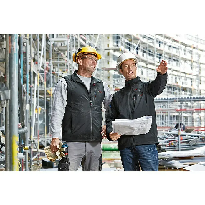 Bosch Professional Arbeitsweste GHV beheizbar - 4 -tlg., Akkuspannung: 12 V, S - Zwei Bauarbeiter, einer mit gelbem Helm und Weste, der andere mit Jacke, diskutieren Baupläne auf einer Baustelle.