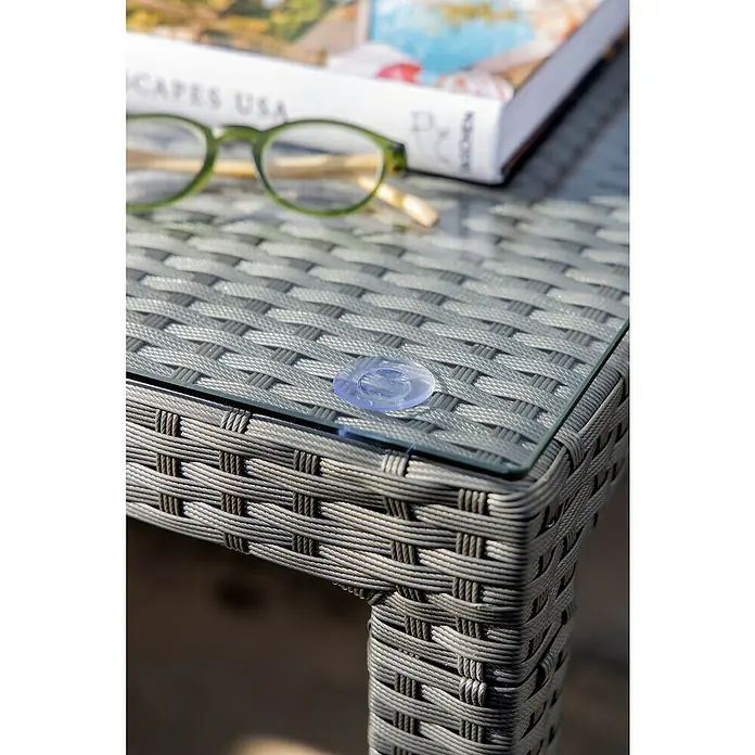Sunfun Mesa de jardín Neila - L x An x Al: 160 x 100 x 75 cm, Aluminio/Ratán sintético, Gris plata - Detalle de una mesa de ratán con tapa de cristal, con gafas y un libro. La textura se destaca por la superficie gris y el diseño trenzado.