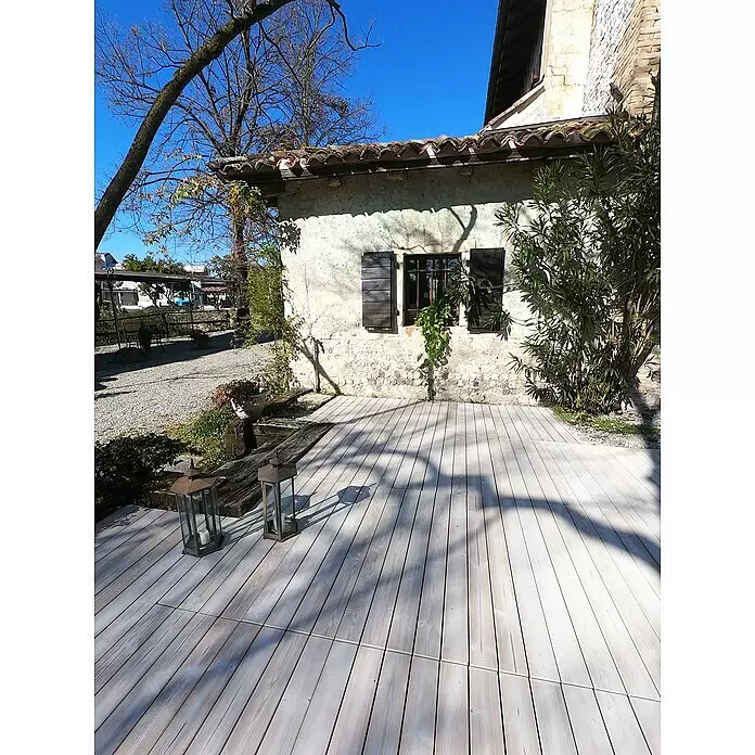 Klickfliese Smartdeck - Kiefer, 120 x 40 x 2,5 cm, Thermo White, 1 Stk. - Hellbraune Holzterrasse vor einem alten Steinhaus, mit zwei dekorativen Laternen darauf.
