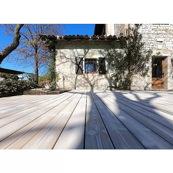 Klickfliese Smartdeck - Kiefer, 120 x 40 x 2,5 cm, Thermo White, 1 Stk. - Hellbraune Holzterrassendielen erstrecken sich im Vordergrund zu einem alten Steinhaus an einem sonnigen Tag.