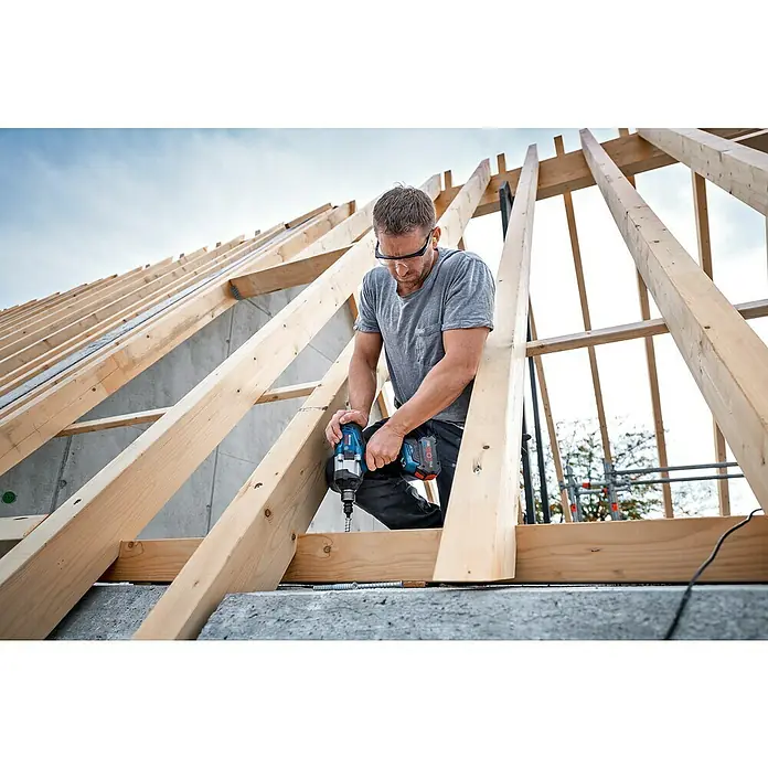 Bosch Professional AMPShare 18V Akku-Drehschlagschrauber GDS 18V-1050 H - 18 V, Ohne Akku - Ein Handwerker arbeitet mit einem Bosch Akku-Schlagschrauber auf einem Dachstuhl, um Holz zu befestigen. Er trägt Schutzbrille für sicheres Arbeiten.