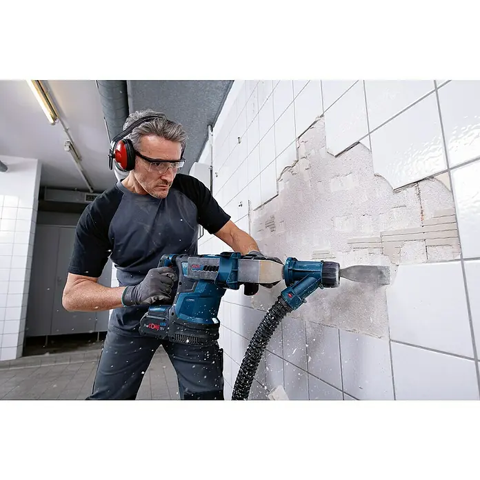 Bosch Professional AMPShare 18V Akku-Kombihammer GBH 18V-34 CF - 18 V, 2 Akkus, 8 Ah, 5,8 J - Ein Mann entfernt Fliesen mit einem blauen Bosch Abbruchhammer, ausgestattet mit einem Absaugschlauch, während er Schutzbrille und Kopfhörer trägt.