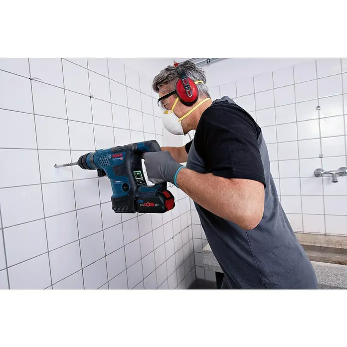 Bosch Professional AMPShare 18V Akku-Kombihammer GBH 18V-34 CF - 18 V, 2 Akkus, 8 Ah, 5,8 J - Ein Handwerker bohrt mit einem blauen Bosch Akku-Bohrhammer ein Loch in eine geflieste Wand, ausgestattet mit Gehörschutz und Schutzmaske.