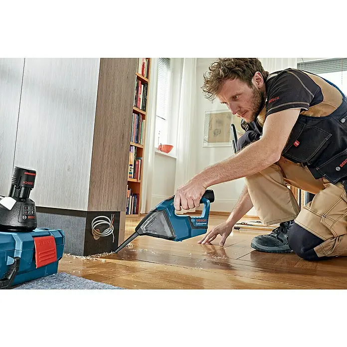 Bosch Professional Akku-Handstaubsauger GAS 12V - 12 V, Ohne Akku, Luftmenge: 15 l/s, Koffer - Ein Handwerker kniet und benutzt einen blauen Bosch-Sauger, um Sägespäne vom Holzboden zu entfernen. Der Bosch-Sauger ist gut sichtbar.