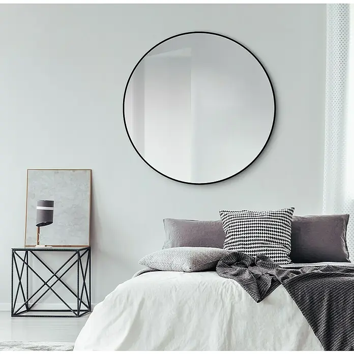 DSK Wandspiegel Silver Globo - Durchmesser: 120 cm, Schwarz - Großes, rundes Spiegelbild mit schwarzem Rahmen über einem Bett mit weißen Laken und grauen Kissen. Dekorative Elemente auf einem Beistelltisch.