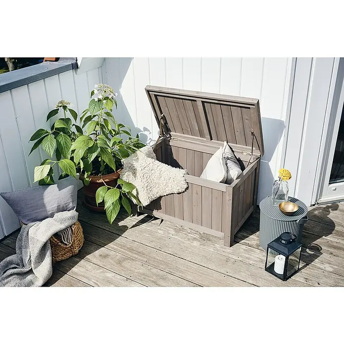 Butenkist Gartenbox Henriette - L x B x H: 75 x 45 x 45 cm, Holz, Warm Grey - Außenaufnahme einer Holzaufbewahrungsbox mit Kissen und Decke, neben einer Pflanze und einem Beistelltisch mit Laterne.
