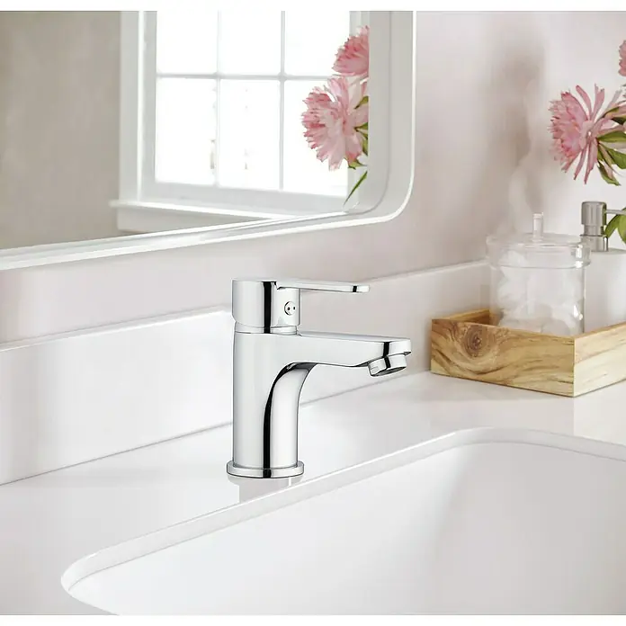 IO Sion Grifo de lavabo - Cromo - Primer plano de un moderno grifo de baño cromado sobre un tocador blanco con espejo y flores decorativas al fondo.