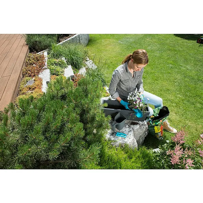 Gardena Gartengeräte - 4 -tlg. - Eine Frau pflanzt Blumen in einen grauen Blumenkasten im Garten, wobei Gartengeräte und ein Sack Blumenerde in der Nähe liegen.