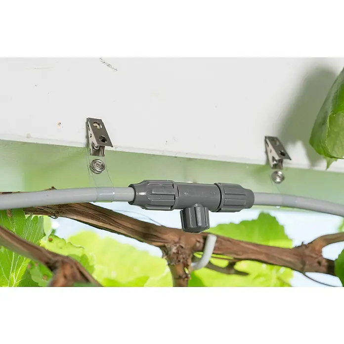 Gardena City Gardening SprühdüsePassend für: Gardena city gardening Outdoor Luftkühler-Set - Nahaufnahme eines grauen Bewässerungsrohrs mit einem T-Stück-Verbinder, das mit Clips an einer weißen Oberfläche befestigt ist und von Pflanzen umgeben ist.