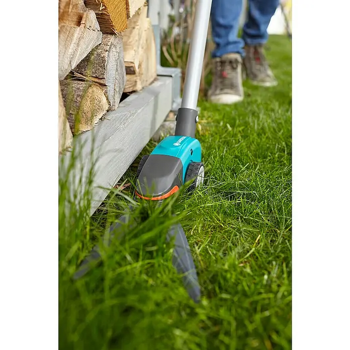 Gardena Comfort Grasschere Langstielig - Bypass, Klingenbeschichtung: Antihaftbeschichtet - Akku-Rasenkantenschere von Gardena beim Trimmen von Gras entlang eines Holzstapels. Komfortable und kabellose Gartenarbeit für präzise Rasenkanten.