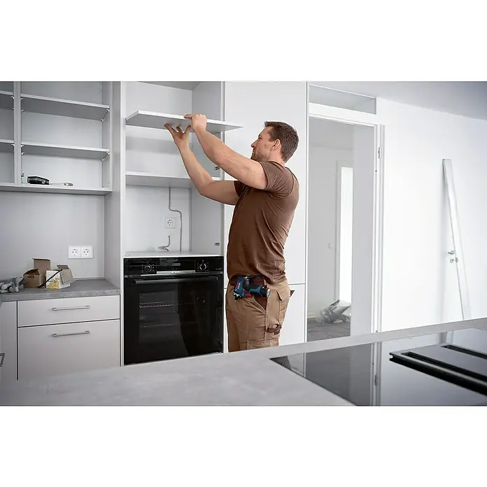 Bosch Professional AMPShare 18V Akku-Bohrschrauber-Set GSR 12V-35 HX PRO SOLO - 12 V, Ohne Akku, 35 Nm, 4 -tlg. - Ein Mann installiert einen weißen Küchenschrank. Der Schrank befindet sich über einem modernen schwarzen Backofen, und eine Bohrmaschine ist am Gürtel des Mannes befestigt.