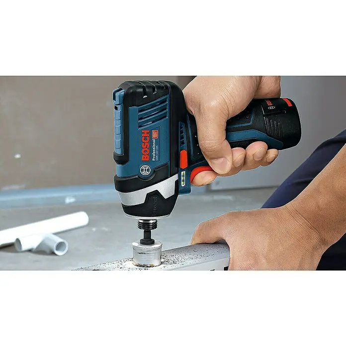Bosch Professional Akku-Drehschlagschrauber - 12 V, Ohne Akku - Ein Bosch Akku-Bohrschrauber beim Bohren in ein Metallstück, mit PVC-Rohren im Hintergrund.