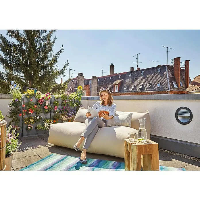 Gardena Blumenkasten-Set NatureUp! Basis Ecke - L x B x H: 17 x 17 x 54 cm - Frau entspannt auf einem beigen Sitzsacksofa auf einer sonnigen Dachterrasse mit bunten Blumen, Wasser und Blick auf die Dächer.