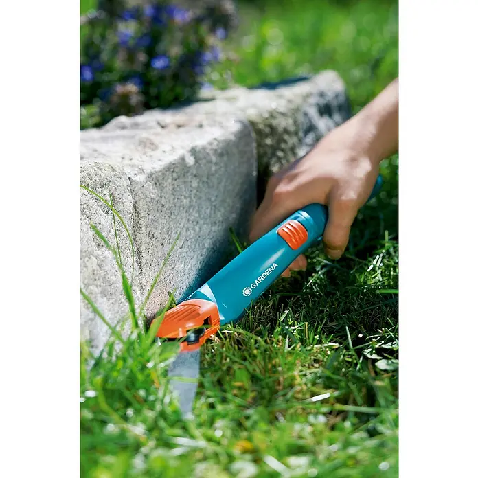 Gardena Grasschere drehbarMaterial Klinge: Geschliffene Schneide - Rasenkantenschere von Gardena im Einsatz, um Rasen entlang einer Steinmauer zu schneiden. Das Werkzeug ist blau und orange.